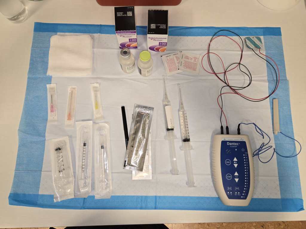 A photo of the countertop of a doctors office showing an EMG machine multiple needles in sealed packaging, multiple needle tips in sealed packaging, two 100-unit vials of Botox and alcohol wipe packets.